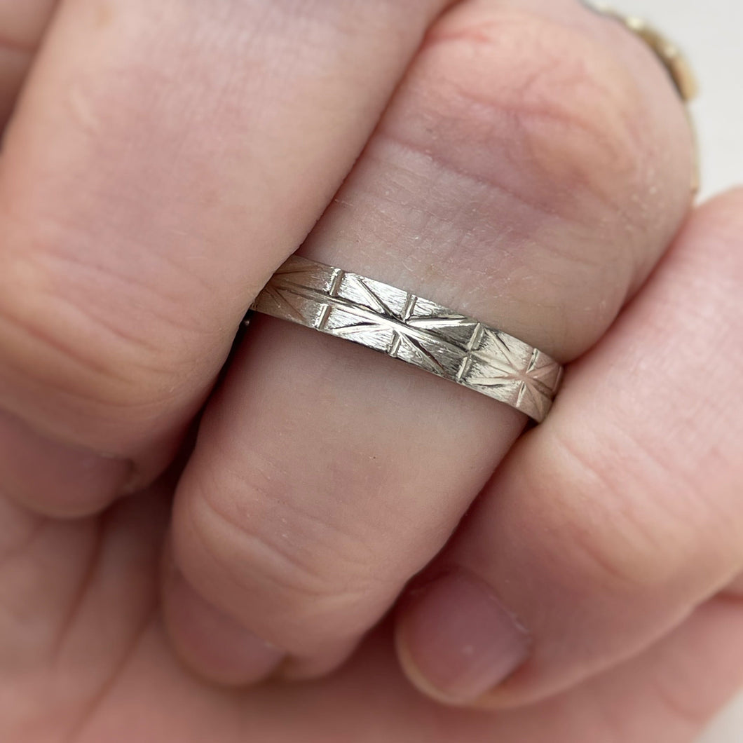 A close-up photo of a white gold wedding band with a geometric-inspired design, handcrafted using ethically sourced and recycled gold. The band measures 4mm wide and 1mm thick and is available in various sizes