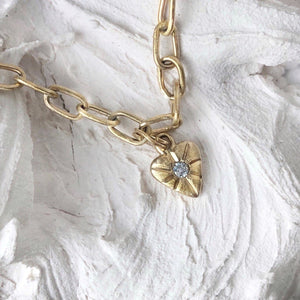 Close-up image of a 14kt yellow gold heart charm with rustic detail and a white sapphire set in the center, attached to two jump rings.
